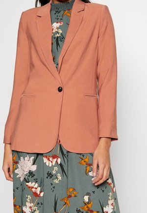 Femme portant un blazer ajusté couleur pêche sur une robe bleue à fleurs avec manches longues et une fermeture à un seul bouton noir.