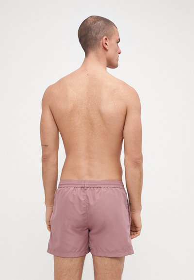 Maillots de bain pour hommes en rose clair, fabriqués à partir d'un tissu doux, dotés d'une taille élastique et de poches latérales, avec une texture lisse et légère.
