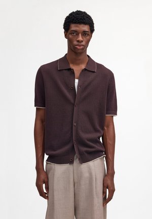 Młody mężczyzna ubrany w ciemnobrązową koszulę z krótkim rękawem zapinaną na guziki, na białej podkoszulce, w jasnobeżowych spodniach, stoi na tle białego tła.