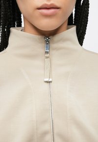 Μπεζ μπουφάν με φερμουάρ και ψηλό γιακά, διαθέτει ασημί μεταλλικό φερμουάρ και απαλό, υφασμάτινο υλικό με υφή. Ο σχεδιασμός περιλαμβάνει ραφές.