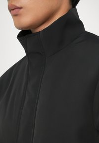 Sort højhalset jakke med en glat, mat tekstur og minimale syninger, der understreger et rent, moderne design.