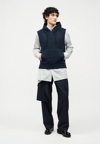 Jonge man met krullend haar, gekleed in een gestructureerde marineblauwe mouwloze hoodie over een lang wit overhemd en zwarte broek met brede pijpen en gelaagde details.