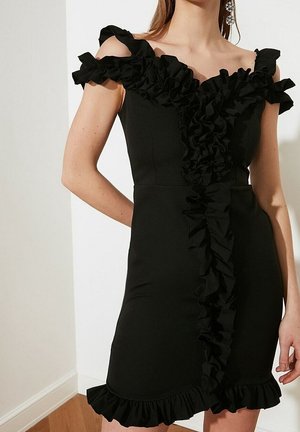 Kobieta ubrana w dopasowaną czarną sukienkę z odkrytymi ramionami, ozdobioną falbanami na ramiączkach oraz pionowym detalem z falbanek z przodu i na dole sukienki.