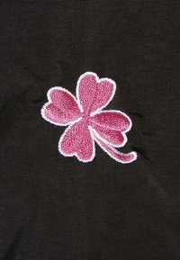 Czarna tkanina z różowym haftowanym wzorem koniczyny, wykończona na biało. Koniczyna ma trzy liście w kształcie serca i zakrzywiony łodygę.