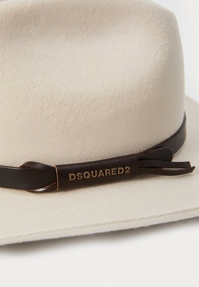Chapéu de feltro bege com uma faixa de couro castanho escuro apresentando um logotipo dourado. A faixa tem borlas decorativas e uma textura lisa.