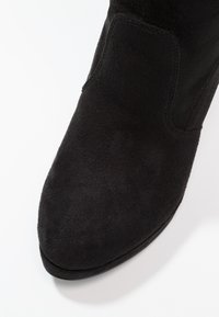 Gros plan sur la pointe d'une botte en daim noir avec des coutures visibles et une forme arrondie légèrement pointue sur fond blanc.