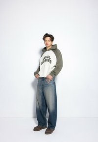 Szary i biały hoodie z napisem "LOCAL HEROES", połączony z luźnymi dżinsami i brązowym obuwiem. Neutralne tło.