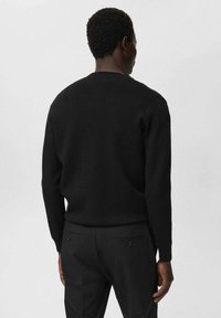 Hombre vestido con una camiseta negra de manga larga y pantalones negros, visto desde atrás contra un fondo claro y liso.