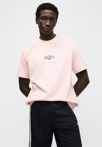 Trumpomis rankovėmis, šviesiai rožinė medvilninė marškinėliai su žaliu "Originals" logotipu. Dėvimi su juodais tinkliniais šortais, pabrėžiantys laisvą siluetą ir raglano rankoves.