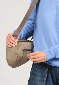 Persoon met een blauwe trui houdt een beige schoudertas vast en maakt de rits van het voorvak los. De tas heeft een verstelbare riem.