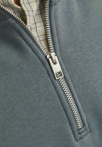 Lys grå zip-sweatshirt med en blød tekstur. Sølv lynlås, der viser et ternet stoffoder under kraven.