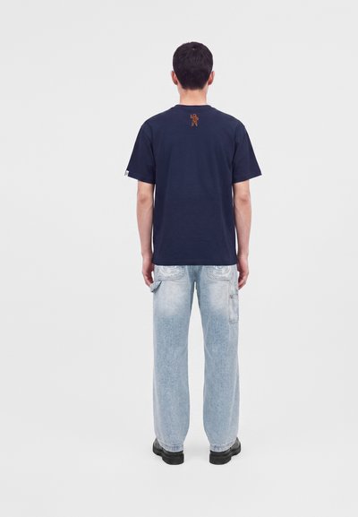 Homme de dos portant un t-shirt bleu marine avec un petit motif astronaute orange, un jean large bleu clair avec des poches et des chaussures noires.