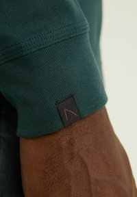 Grön bomullssleeve med ribbat mönster, försett med en svart etikett och orangea sömdetaljer. Slät yta och kroppsnära passform.