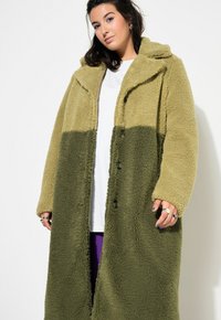 Cappotto sherpa verde e beige con cappuccio, caratterizzato da chiusura a bottoni, design bicolore e tessuto strutturato, che arriva a metà polpaccio.
