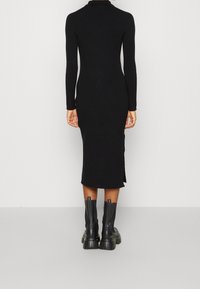 Robe en tricot côtelé noire avec une silhouette ajustée, des manches longues, un col haut et une fente sur le côté, associée à des bottines épaisses noires.