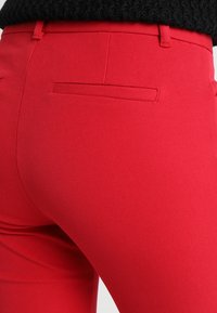 Close-up van een persoon die een strakke, felrode broek draagt met een achterzak en een zwart gebreid topje.