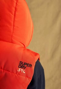 Gilet rembourré rouge avec capuche, fabriqué en tissu lisse. Présente des logos brodés en noir "SUPER DRY" et "JPN." sur l'épaule.