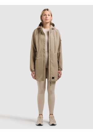 Woman standing wearing a beige hooded jacket over matching leggings and sneakers, facing forward against a plain gray background.