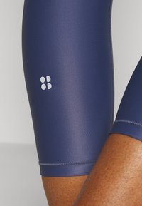 Primer plano de una persona que lleva pantalones cortos de ciclismo ajustados, de color azul oscuro, con un pequeño logo blanco de cuatro puntos en el muslo.