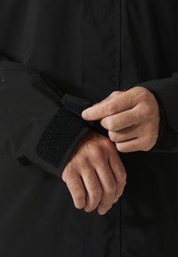 Zwarte waterdichte jas met klittenbandsluiting bij de manchetten, met een gladde textuur en ergonomische pasvorm voor comfort en bescherming.