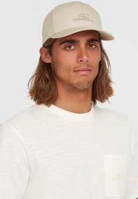 Beige kasket med en buet skygge og broderet logo. Båret med en lysefarvet, tekstureret skjorte med en brystlomme.