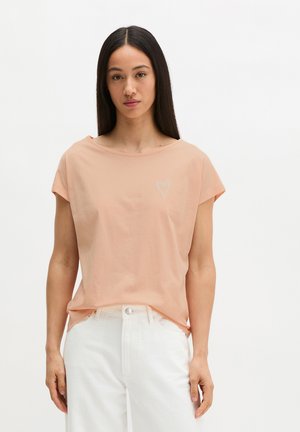 Junge Frau mit langen dunklen Haaren, die ein pfirsichfarbenes kurzärmliges Oberteil mit kleinem Herz-Detail und weiße hoch taillierte Hosen trägt.