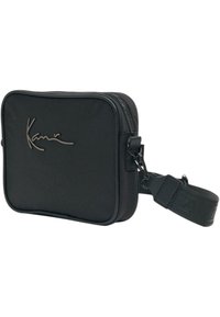 Czarna prostokątna torba wykonana z materiału syntetycznego, z zamkiem błyskawicznym z przodu, regulowanym paskiem oraz nazwą marki wytłoczoną z przodu.