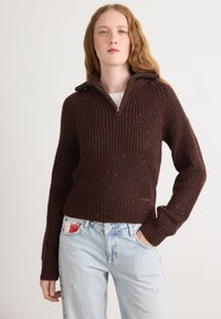 Bruine geribde trui met een halve ritssluiting, een gestructureerd patroon en kleurvlekken doorheen. Draag met lichtblauwe distressed jeans.