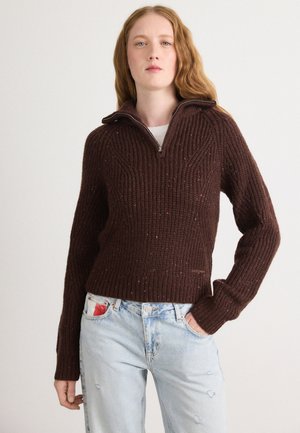 Bruine geribde trui met een halve ritssluiting, een gestructureerd patroon en kleurvlekken doorheen. Draag met lichtblauwe distressed jeans.
