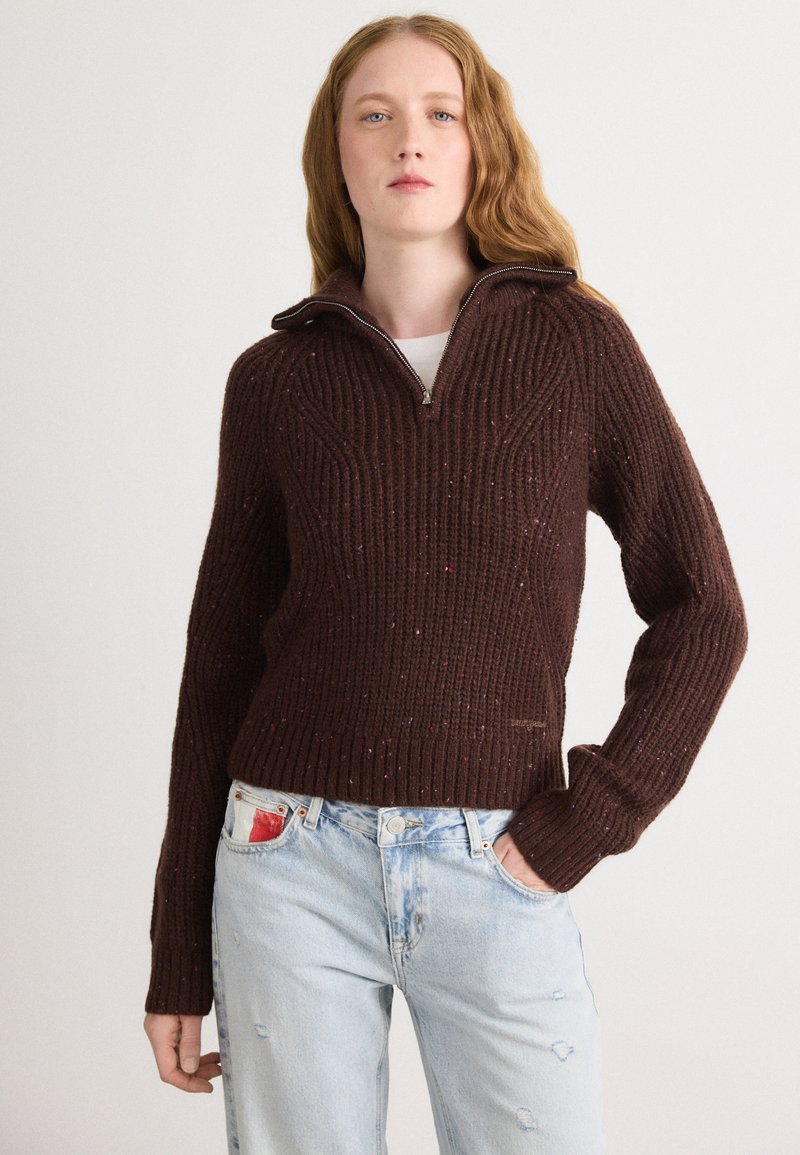 Bruine geribde trui met een halve ritssluiting, een gestructureerd patroon en kleurvlekken doorheen. Draag met lichtblauwe distressed jeans.