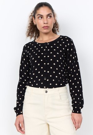 Woman with curly hair wearing a black long-sleeve polka dot cardigan and high-waisted white pants looking to the side.