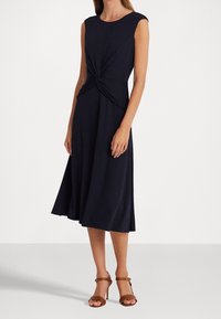 Marinblå ärmlös klänning med knytdesign vid midjan, A-linjeformad kjol och mjukt material, kombinerad med bruna högklackade sandaler.