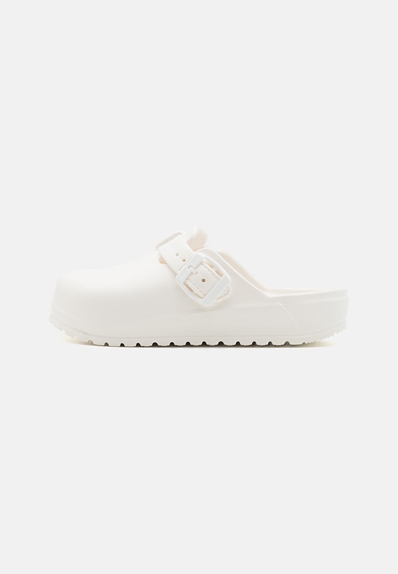 Sabot blanc à enfiler avec boucle latérale réglable et semelle texturée, présenté de profil sur fond uni.