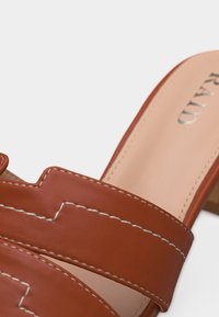 Sandale en cuir marron à bout ouvert avec des coutures blanches et un talon carré sur un fond clair.