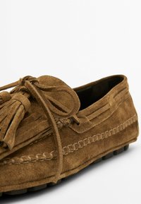 Brun ruskindsløs med kvast og sløjfedetalje, mokassinsyning og sål med gummigreb, vinklet for at vise side og front.