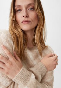 Jasno beżowy sweter dzianinowy o puszystej fakturze, z prążkowanymi mankietami i luźnym krojem, noszony przez modelkę z długimi, falowanymi włosami.