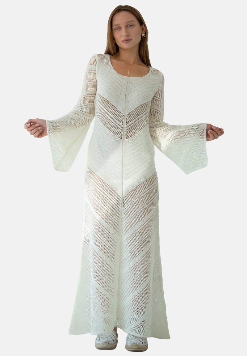 Długa, biała, dziana sukienka z wzorem w zygzak, z dekoltem łódkowym, rozkloszowanymi rękawami i teksturowanym wykończeniem, sięgająca do połowy łydki. Noszona z butami wsuwanymi.
