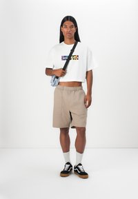 Hvit kortermet T-skjorte med flerfargede "CONVERSE"-logo, beige shorts med sidelommer, grå skulderveske, svarte joggesko med hvite detaljer.