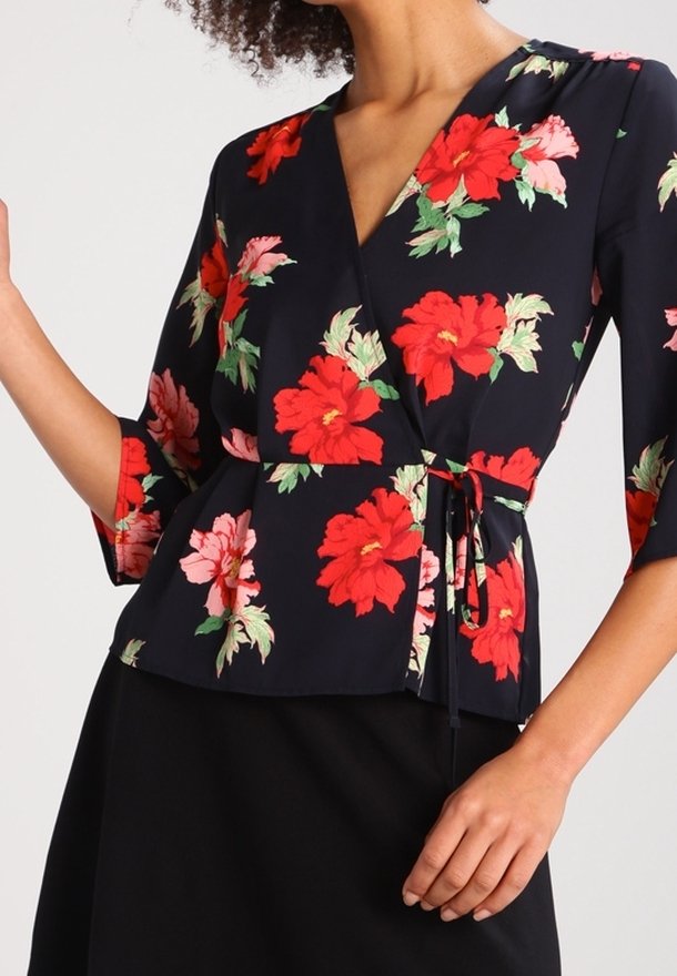 Haut cache-cœur fleuri avec manches trois-quarts. Fond bleu marine avec des fleurs rouges et roses. Ceinture nouée à la taille. Tissu léger.