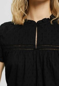Blouse noire en tissu texturé présentant une encolure clé avec boutons, des manches courtes et de petits motifs brodés sur l'ensemble.