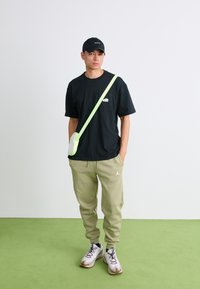 Ung mand iført sort kasket og t-shirt, beige sweatpants, hvide sneakers og en neon grøn crossover-taske står på en grøn gulv.