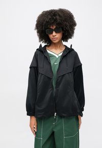 Μαύρο τζάκετ με φερμουάρ και κουκούλα, κατασκευασμένο από ελαφρύ υλικό. Διαθέτει ελαστικά μανίκια και μοναδικό στρωματοποιημένο σχέδιο. Φορεμένο πάνω από πράσινο αθλητικό τοπ.