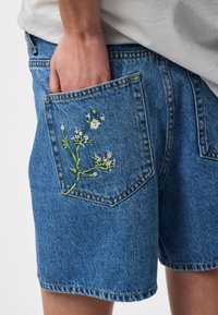 Main dans la poche arrière d'un short en denim bleu avec broderie florale blanche, associée à une chemise blanche.