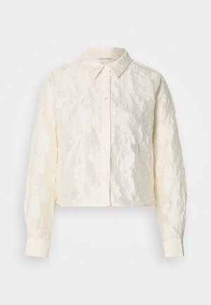 Μπεζ κοντή πουκάμισα με υφασμάτινη υφή, κουμπιά στο μπροστινό μέρος και μακριά μανίκια. Διαθέτει κλασικό γιακά και ελαφρώς ρυτιδωμένη εμφάνιση.