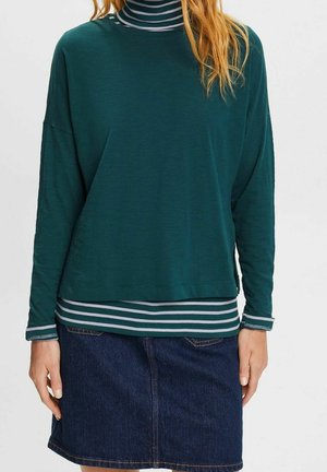 Kvinde iført en mørkegrøn langærmet top over en grøn og hvid stribet rullekrave med en mørkeblå denimnederdel.