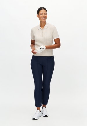 Beige gebreid poloshirt met korte mouwen, marineblauwe slim-fit broek en wit golfhandschoen. Witte sneakers met zwarte zolen maken de outfit compleet.