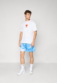 Fehér pamut póló narancssárga grafikával és fekete Nike logóval, kék tye-dye rövidnadrággal, fehér zoknival és fehér sportcipővel párosítva.