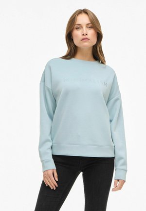 Mujer vestida con un sudadera ligermente azul que lleva el texto "MINIMALISMO" y jeans negros, de pie frente a un fondo blanco liso.