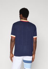 Morska plava rebrasta pamučna t-shirt s ovratnikom i rukavima u crvenoj, žutoj i bijeloj boji. Klasičan kroj s kratkim rukavima.
