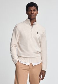 Maglione zip a coste di colore beige chiaro realizzato in morbido materiale a maglia, con polsini e orlo a coste, e un piccolo logo scuro sul petto.